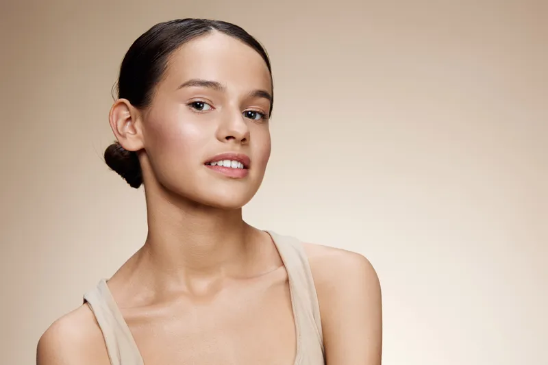 Natural beauty portrait of a young woman with soft features, wearing neutral makeup and a simple tank top, set against a warm beige background