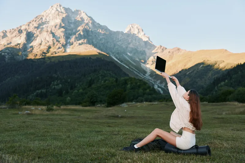 A young woman sits on a grassy field, raising a laptop toward the sky as rugged mountains rise behind her, creating a playful, techy outdoor moment of exploration and focus.