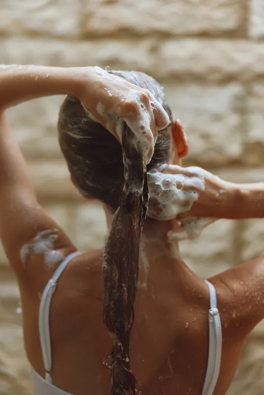 Woman washing long hair with shampoo foam in shower, wet brown hair, bare shoulders, and white straps visible, natural stone wall background creating a warm atmosphere.