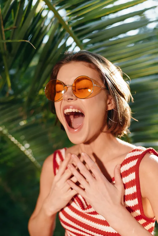 A joyful woman wearing round orange sunglasses and a red striped top reacts with a wide open mouth in a sunny outdoor setting, expressing excitement and carefree energy.