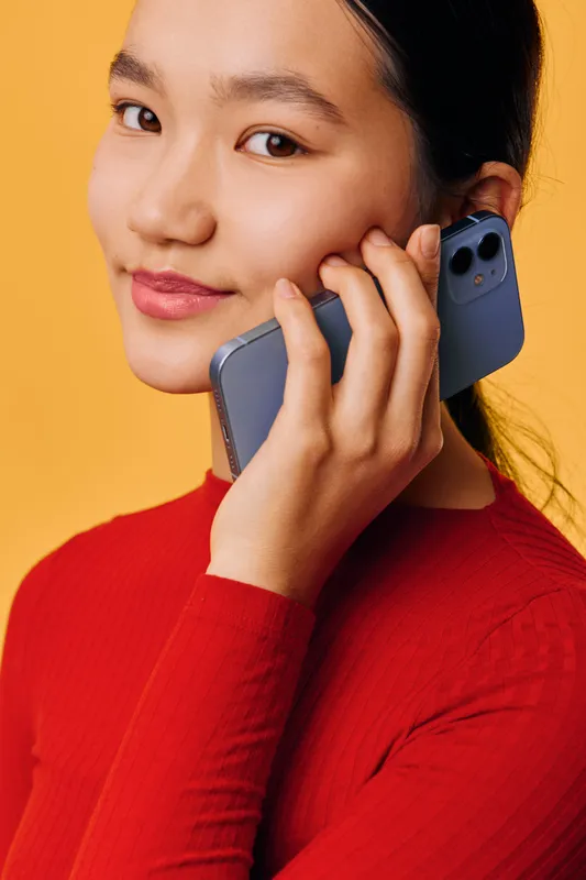 Young woman in red sweater holding smartphone to ear with smile on yellow background. Portrait of happy female using mobile phone for communication and casual call.