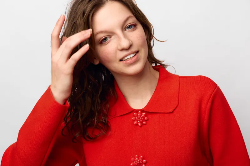 Young woman with curly brown hair wearing a red sweater smiles gently while touching her hair against a plain white background. Concept of natural beauty and casual style.