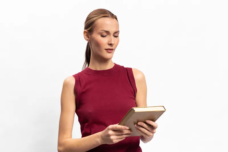 Young woman with light skin and pulled back hair wearing red sleeveless top reading a book against white background. Concept of education, knowledge, and focused learning.