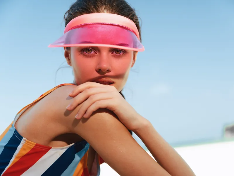 Bright portrait of a young woman wearing a pink visor and a striped swimsuit, resting her chin on folded arms under a clear blue sky, radiating playful confidence and summer energy.