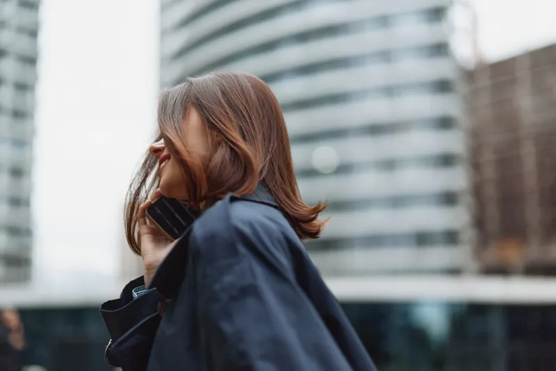 Young woman with brown hair talks on a mobile phone in a city street, wearing a navy coat, capturing a candid urban moment and contemporary life.