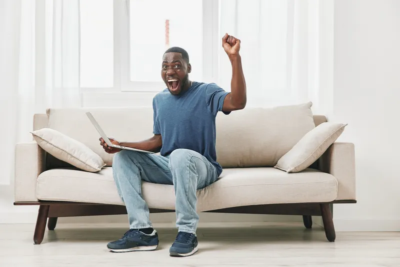 Happy man celebrating success while sitting on a couch with a laptop, expressing joy and excitement in a bright, modern living room