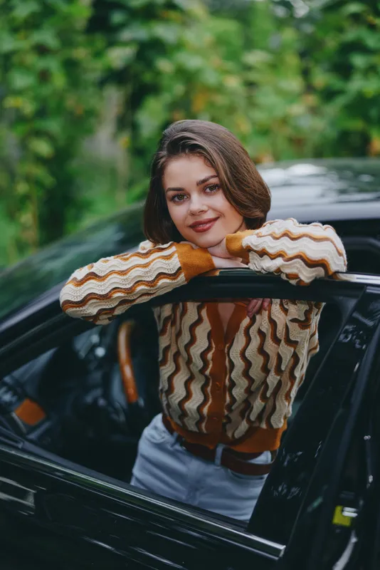 Young woman leaning on a car window, wearing a striped knit sweater, captured in natural daylight with a relaxed, confident expression.