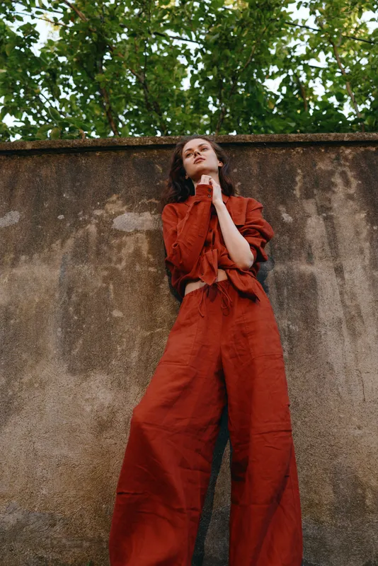 Woman wearing red overalls stands against a textured wall outdoors, relaxed pose and thoughtful gaze, conveying casual urban lifestyle with natural light and moody atmosphere