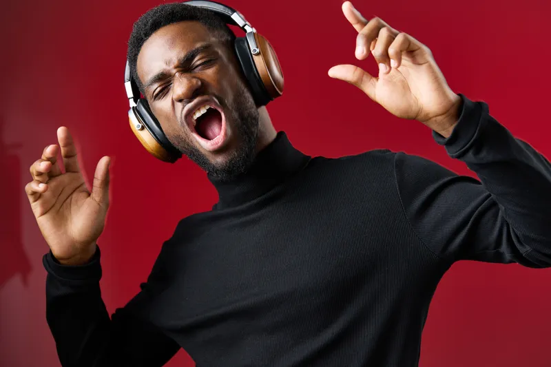 Young African man enjoying music with headphones, singing passionately against a vibrant red background, showcasing a fun and energetic vibe