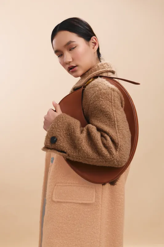 asian woman wearing a camel coat with a round brown bag slung over her shoulder, standing in a minimal studio with soft lighting and a calm, poised expression.
