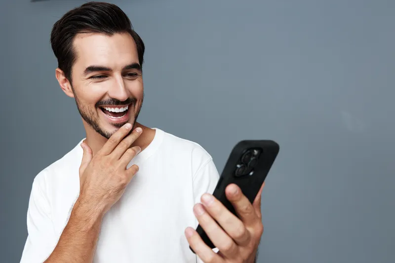 Smiling happy cheerful young man wearing a casual white t-shirt, looking at his mobile phone with excitement, isolated on a plain gray background. People lifestyle concept.