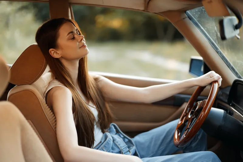 Young woman enjoying a peaceful moment in a vintage car, with sunlit nature in the background, wearing casual blue jeans and a relaxed expression, evoking freedom and serenity.