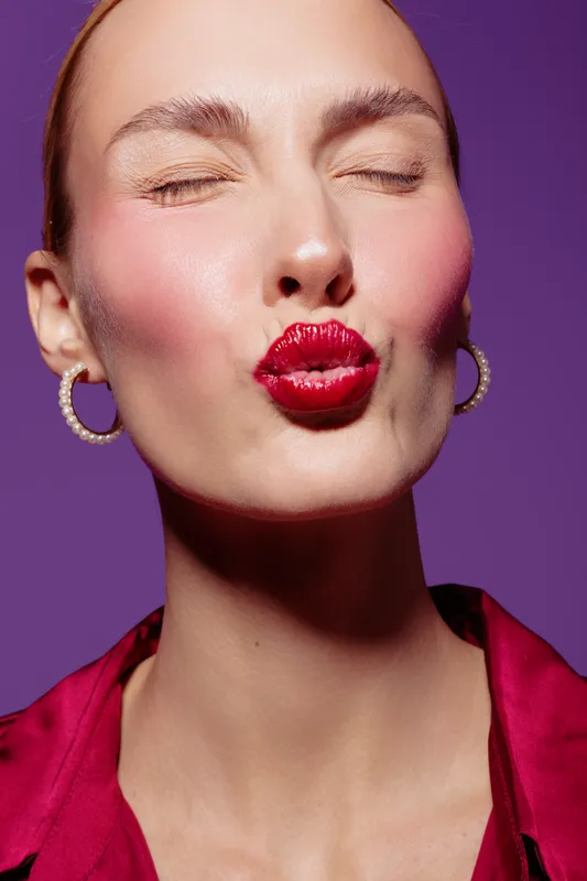 Close-up of a woman with red lipstick making a kiss face, eyes closed, wearing hoop earrings and a red blouse. Studio portrait on purple background with bright makeup and smooth skin.