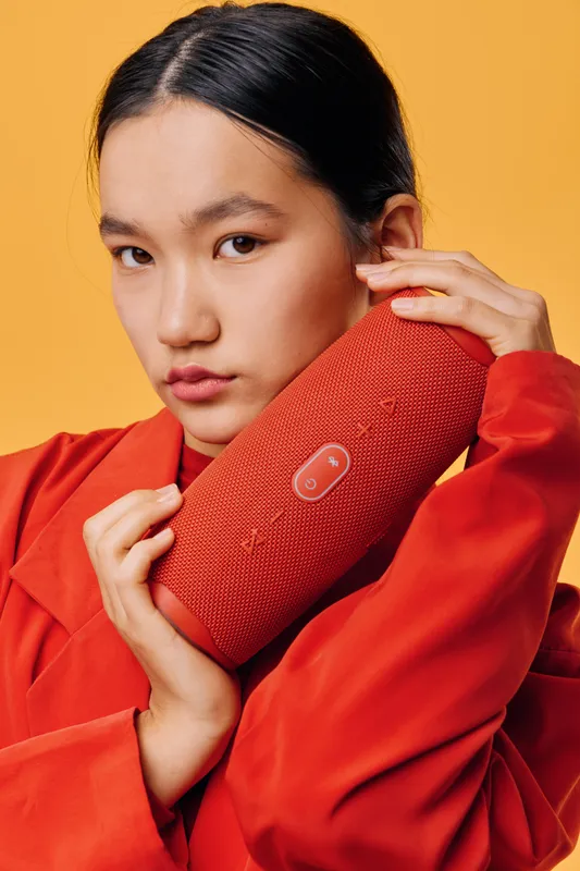 Young woman in red outfit holding portable wireless speaker close to face against orange background. Asian girl with serious expression posing with audio device, fashion and technology concept.