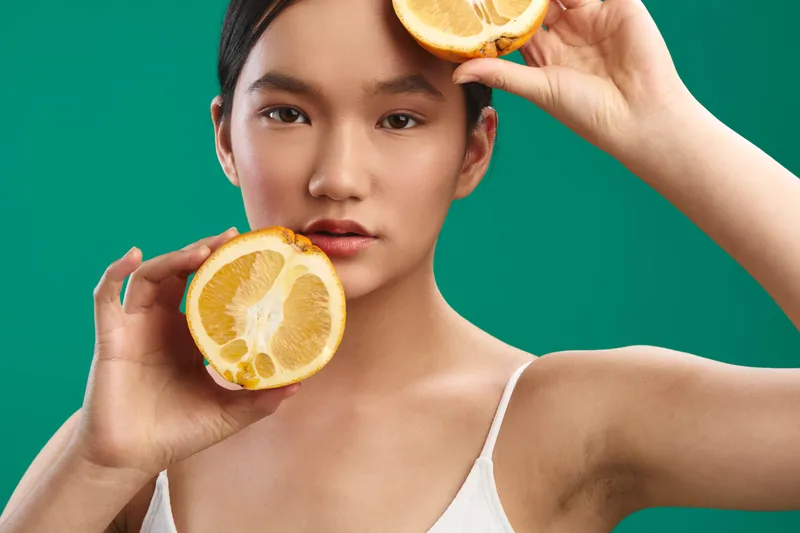 asian woman poses with citrus slices near her face in a vibrant studio, expressing freshness and confidence, inviting healthy lifestyle and modern beauty moments.