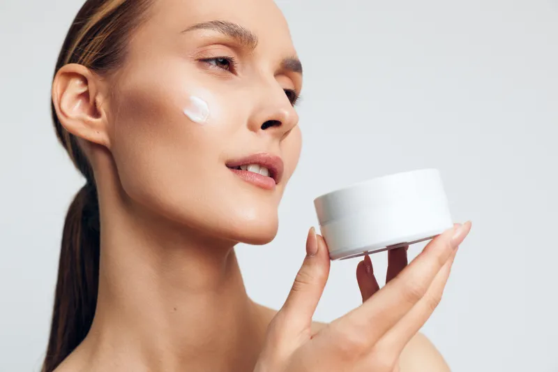 Young woman with clear skin applying white face cream on cheek holding open container. Skincare concept with natural beauty and wellness on plain light background. Soft lighting highlights smooth