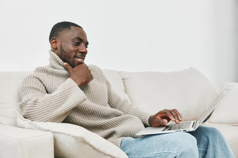 Young African man in cozy sweater enjoying online browsing on a laptop in a modern living room, showcasing relaxed and content emotions