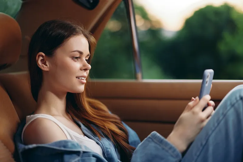 Young woman in casual denim outfit using smartphone in a car, smiling with relaxed mood, surrounded by green nature, showcasing modern lifestyle and technology.