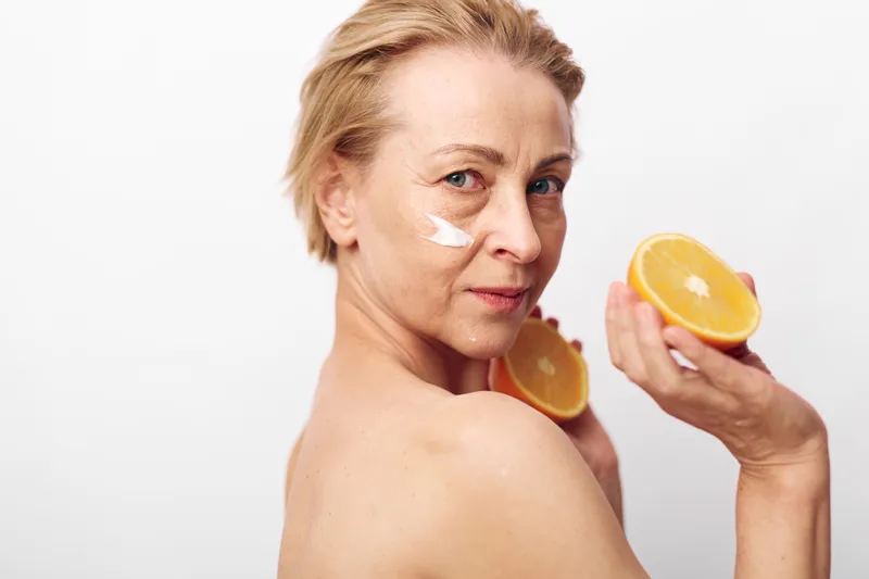 Mature woman with clear skin applies white face cream on cheek holding fresh orange halves. Concept of natural skincare, healthy lifestyle, organic beauty and facial care routine.