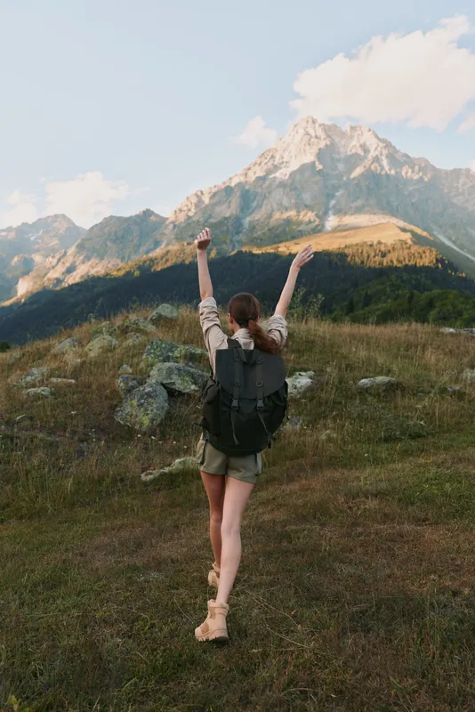 Smiling woman with a backpack raising her arms in a grassy meadow, mountains in the background. Outdoor lifestyle, adventure, and personal achievement. Bright natural light.