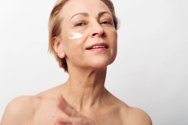 Mature woman with short hair applies white face cream on cheek, smiling gently. Close-up on light background. Concept of skincare, aging beauty, self-care, and natural wellness.