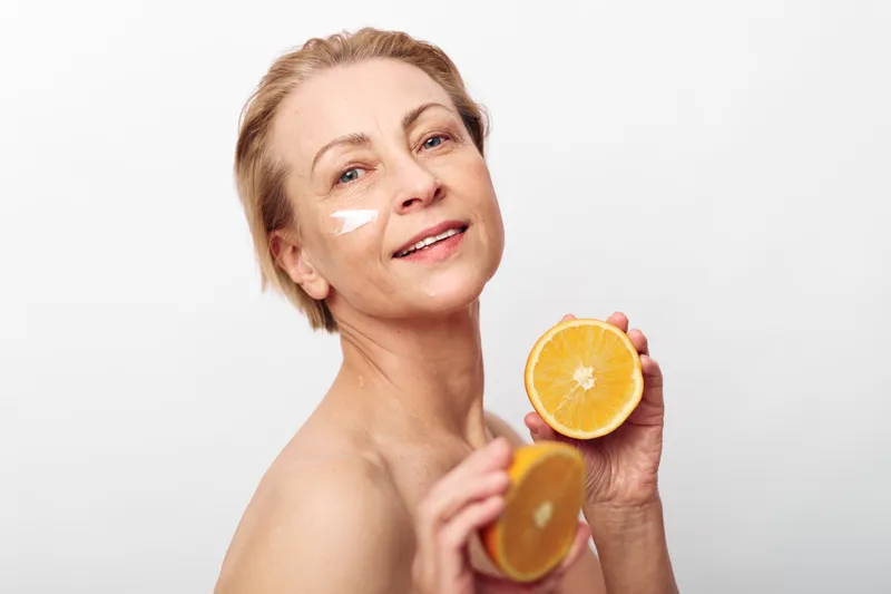Mature woman with natural skin care cream on face holds two halves of fresh orange, smiling gently against white background. Concept for healthy lifestyle, vitamin C, and beauty routine.