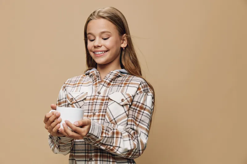 Girl smiling and holding white cup with both hands wearing plaid shirt on beige background. Happy child enjoying warm beverage with eyes closed and relaxed pose.