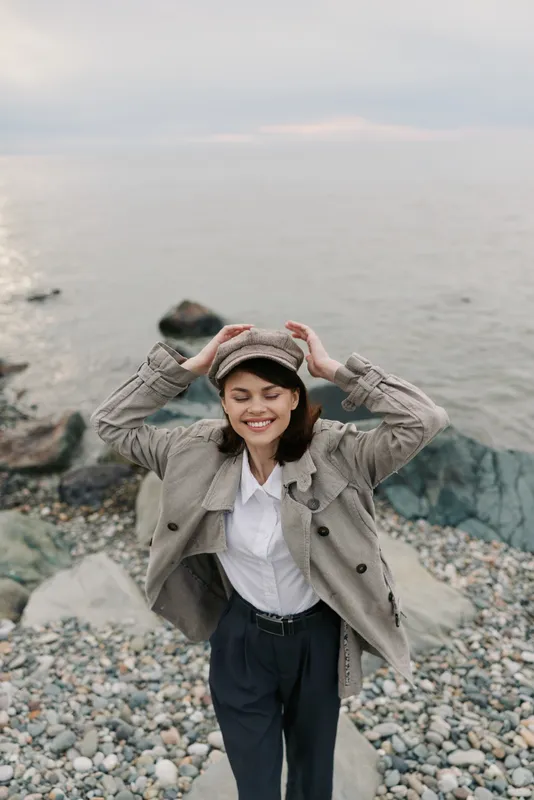 Smiling woman in a plaid coat and hat enjoys a windy day on a rocky beach, eyes closed and hands adjusting hat. Concept of autumn fashion, nature, and happiness outdoors.