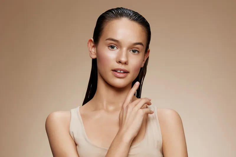 Young model with wet hair, wearing a nude top, poses gracefully against a soft beige background, expressing calm confidence and natural beauty