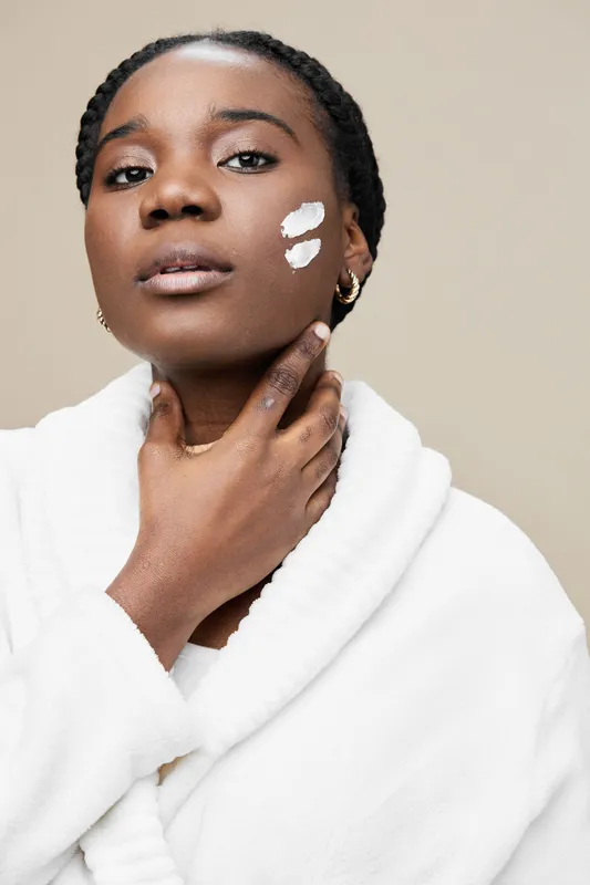 Portrait of a young Black woman applying skincare cream, showcasing natural beauty and self care in a minimalistic setting with a neutral background
