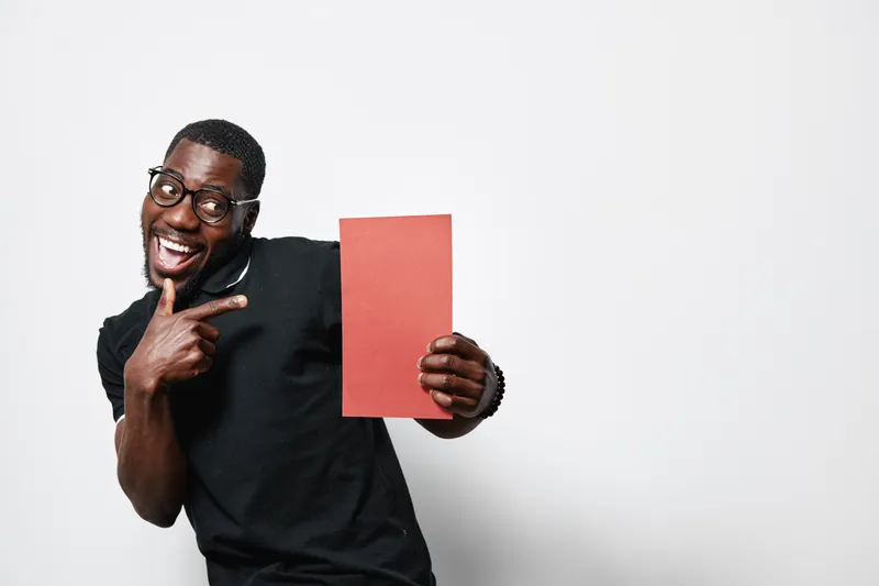 African American man with dark skin and glasses holding red paper and smiling while pointing at it on white background. Casual clothing and happy expression with positive gesture.