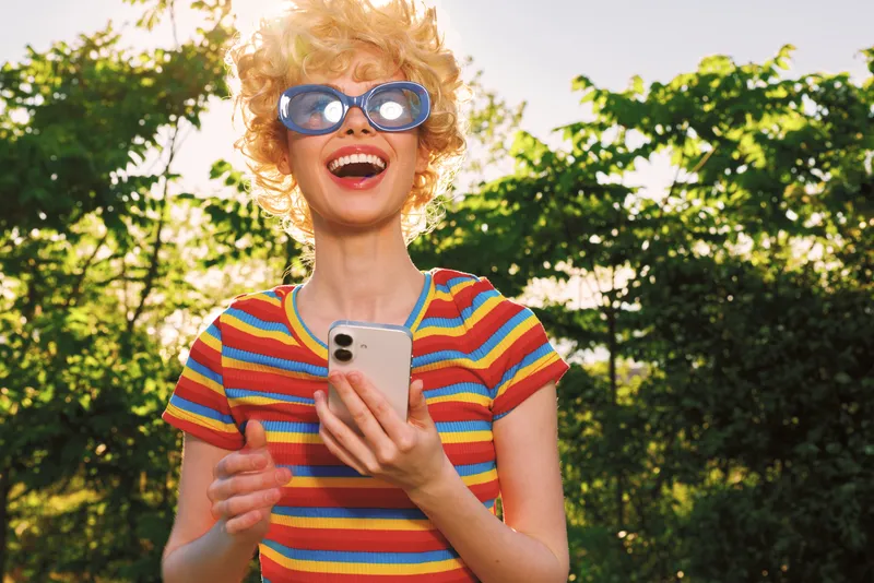 Young woman in a colorful striped shirt laughs outdoors, holding a smartphone as sunlight highlights her carefree mood.