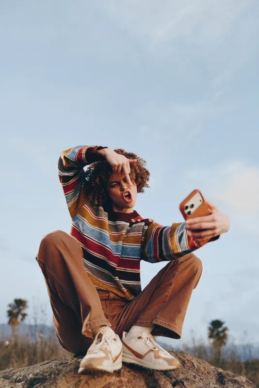 Outdoor lifestyle, woman taking selfie with smartphone, sitting on rock, striped sweater and jeans, relaxed calm mood, summer sunlight, blue sky and distant trees behind her, soft clouds nearby