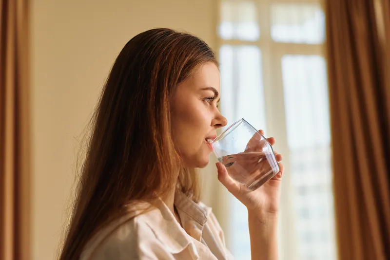 Woman drinking water from glass in profile indoors by window, relaxed morning routine and hydration for wellbeing, natural light on face and casual shirt and calm expression.