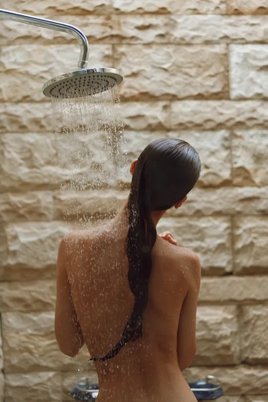 woman showering outdoors under rain showerhead with wet hair against stone wall background in natural light, showing relaxation and freshness