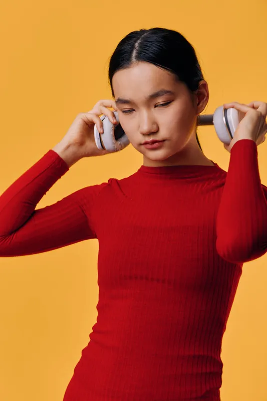 Young woman in red sweater wearing wireless headphones with eyes closed on orange background, enjoying music and relaxing with modern technology indoors.