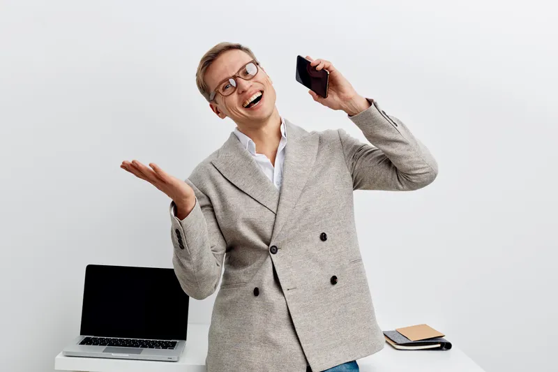 Smiling woman in business casual attire holding smartphone, standing at modern office desk with open laptop and notebook, expressing happiness and success, white background, studio shot, people lifest