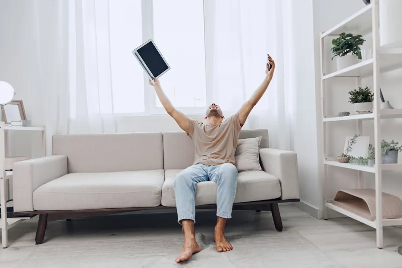 Happy man celebrating success at home, raising a laptop and smartphone. Bright, minimalistic interior creates a positive, uplifting atmosphere for modern lifestyles.