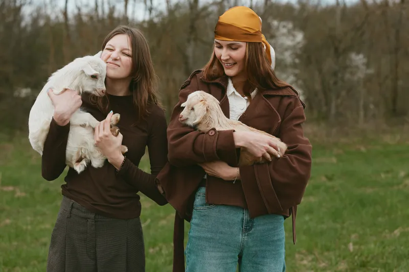 A candid lifestyle shot shows two women outdoors with small goats, smiling, holding playful kids, illustrating farm life, companionship, and gentle animal interaction in a grassy field, with natural