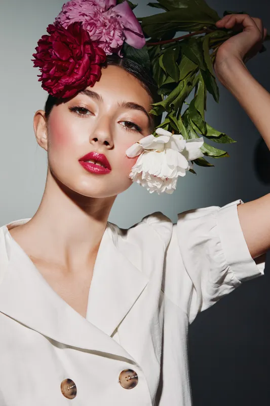 Beautiful young woman with bright pink and red flowers styled in her hair looks confidently at camera in white outfit. Concept of beauty, creative fashion, and floral art.