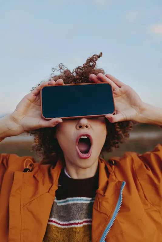 Woman with curly hair outdoors, holding a smartphone over her eyes, surprised open mouth, orange jacket and striped sweater, hands framing the screen, blue sky background, lifestyle