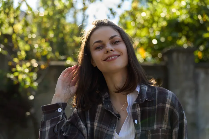 Young woman outdoors wearing a plaid shirt smiles softly as warm sunlight filters through green foliage, creating a relaxed, natural mood and casual charm