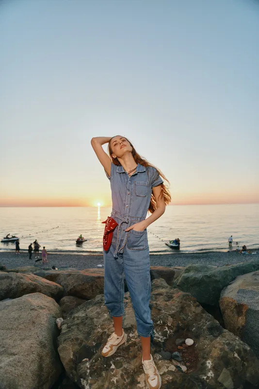 lifestyle woman in modern boho-western denim street style poses confidently on rocky beach during sunset, film color capturing relaxed summer vibe and casual fashion.