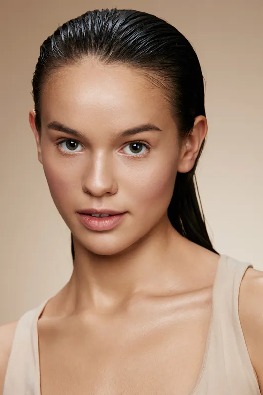 A fresh faced young woman with wet hair and minimal makeup, radiating natural beauty and confidence against a neutral beige background