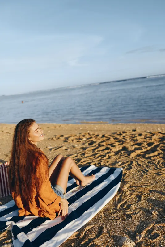 woman beach ocean sand sunlight summer relaxation blanket outdoors coast shore horizon calm waves soft clouds serene pose seated with smile natural colors warm golden light on the sand