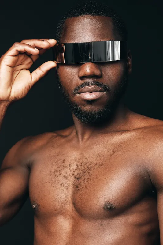 Black man with bare chest wearing futuristic silver visor in creative conceptual visual style. Portrait of African ethnicity with modern metallic eyewear on dark background for innovation and fashion.