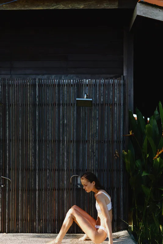 A woman in a swimsuit sits beside an outdoor shower, dark wall behind. Sunlight glistens on water droplets, creating a relaxed lifestyle moment focused on solitude and warm refreshment.