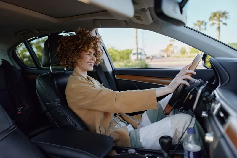 Curly haired woman driving a car with a relaxed smile, casual chic outfit, warm retro film color, daylight inside the cabin, sense of freedom and everyday travel.
