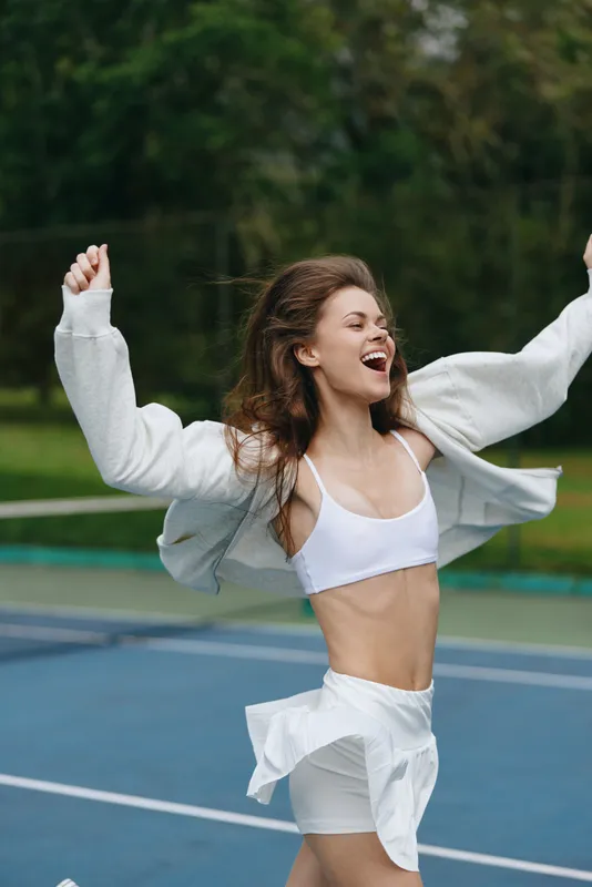 Joyful young woman celebrating on a tennis court in sporty attire, showcasing a healthy lifestyle and vibrant energy in an outdoor setting