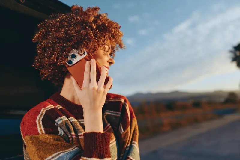 Woman with curly hair holding a smartphone to her ear outdoors, wearing a striped sweater, side profile with relaxed smile, warm sunlight, lifestyle street scene with sky and landscape